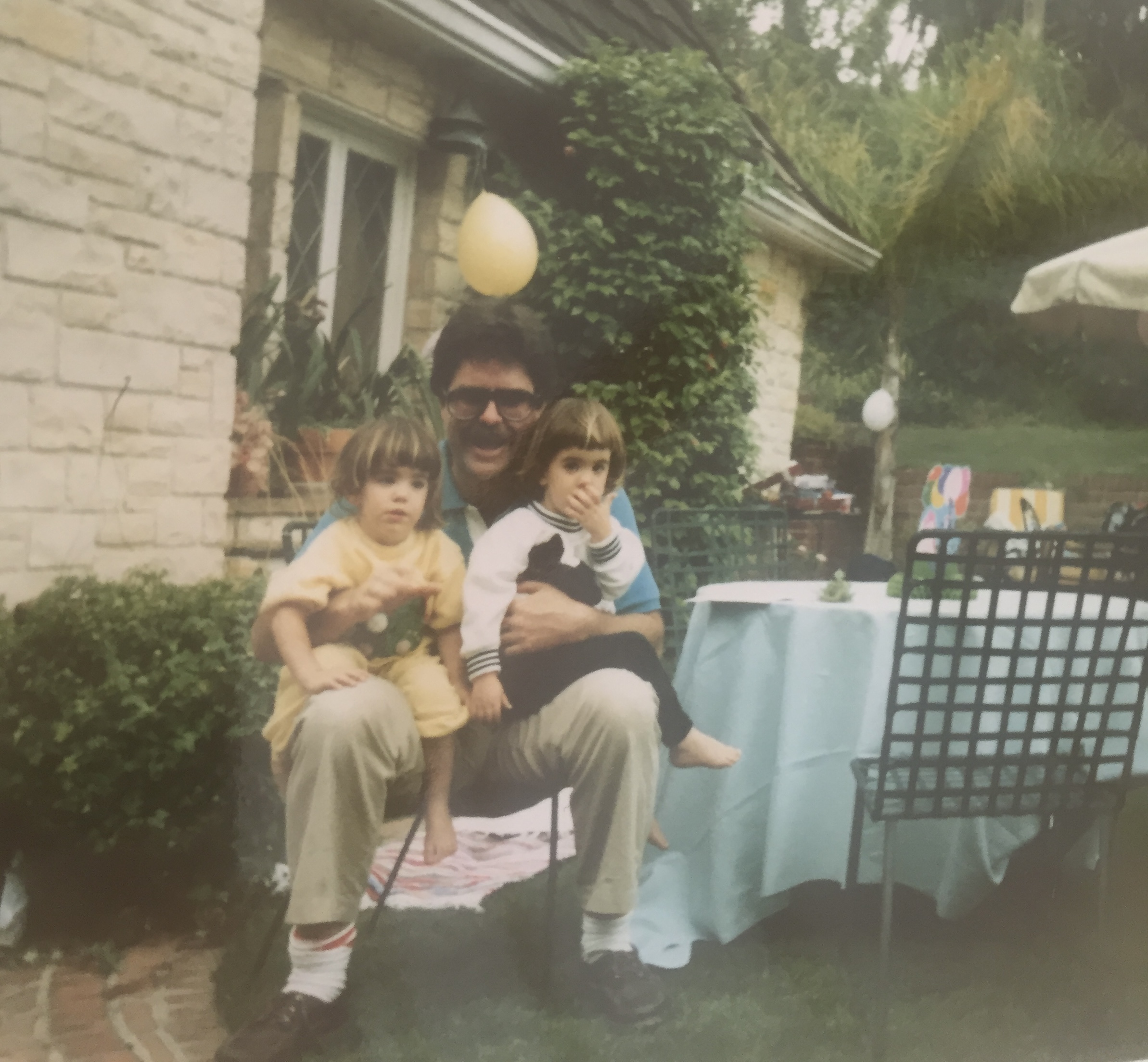 A joyful family moment in a sunlit garden—two children sit close with their parent, surrounded by colorful balloons, lush greenery, and the rustic charm of a stone house. The scene radiates warmth, connection, and the simple beauty of being together.