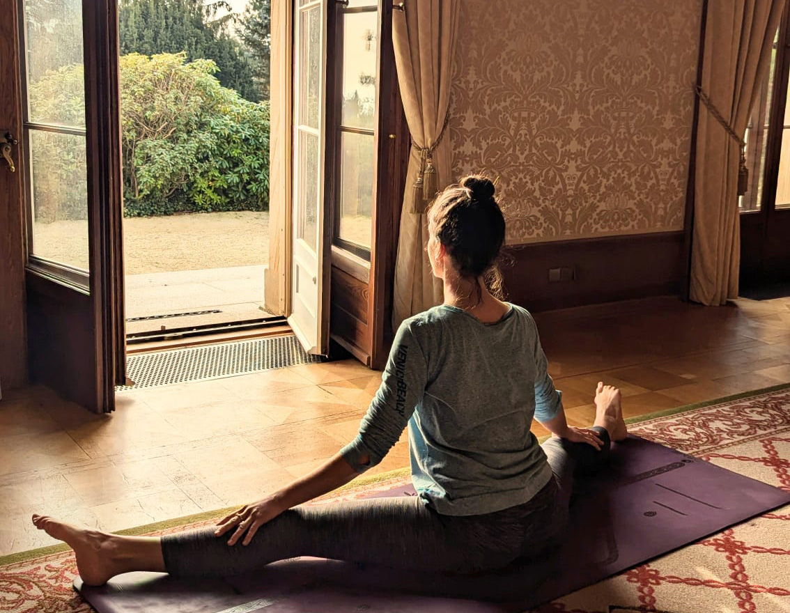 A serene moment of self-connection—Katie sits in a gentle yoga pose on a mat, bathed in natural light from open doors that lead to a lush garden. The room’s soft textures and earthy tones create a peaceful sanctuary, inviting stillness, breath, and quiet reflection