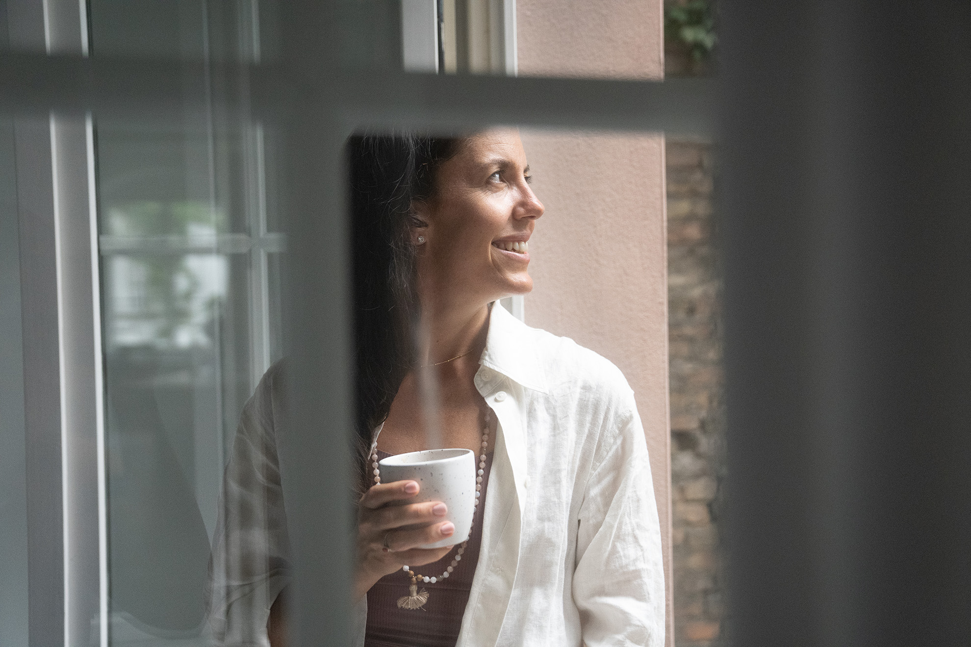 A yoga instructor stands by a sunlit window, holding a cup in quiet reflection—radiating calm, presence, and everyday beauty.